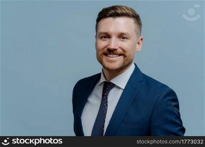 Satisfied cheerful male entrepreneur or business man wearing classic suit smiling happily at camera while standing isolated over light steel blue background. Career growth and success concept. Portrait of happy handsome business man smiling at camera