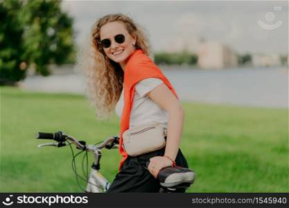 Satisfied curly female bicyclist enjoys spare time, travels on bike, stops to have little rest, wears sunglasses, casual summer outfit, poses among grass and trees, green blurred background.