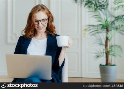 Satisfied European female with ginger hair, types information on laptop computer, has glad facial expression, drinks coffee, wears spectacles, develops new project, poses in cosy room alone.