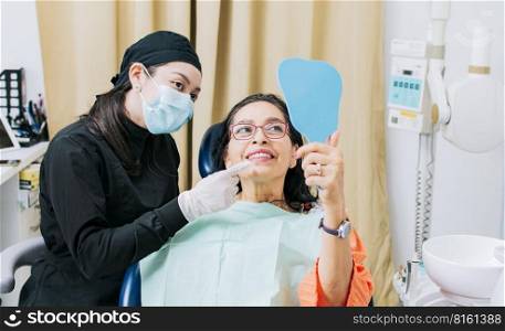 Satisfied female client in dental clinic looking at mirror, Dentist with patient smiling at hand mirror in office, female patient checking teeth after curing teeth in dental clinic,