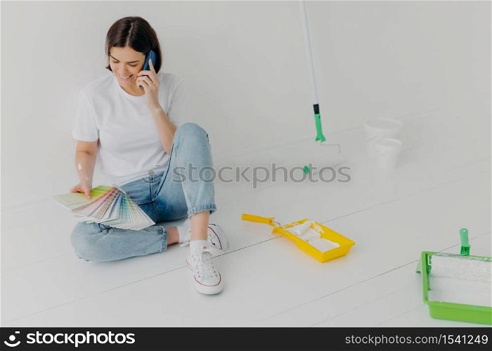 Satisfied female designer discusses ideas via smartphone, looks attentively at color samples, sits on floor in empty room which needs redecoration, has cheerful mood, chooses colour for painting