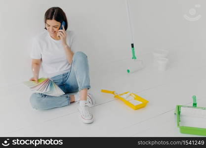 Satisfied female designer discusses ideas via smartphone, looks attentively at color samples, sits on floor in empty room which needs redecoration, has cheerful mood, chooses colour for painting