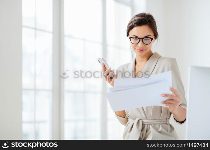 Satisfied female employee focused in documents, reads prepared report attentively, holds modern smartphone, wears transparent glasses and business suit poses over office interior checks monthly income