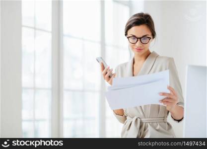 Satisfied female employee focused in documents, reads prepared report attentively, holds modern smartphone, wears transparent glasses and business suit poses over office interior checks monthly income