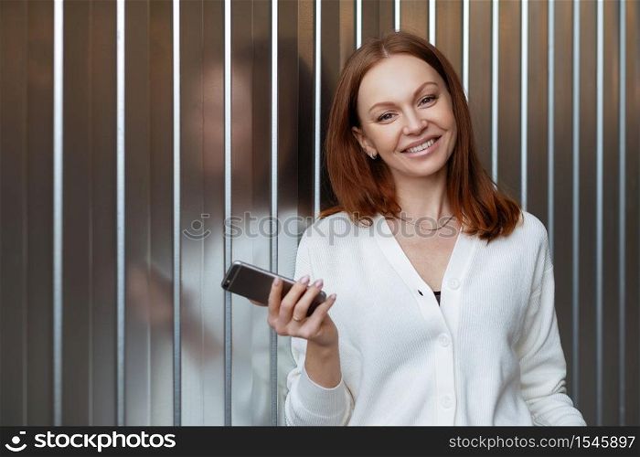 Satisfied female entrepeneur with toothy smile, wears white jumper, holds modern cell phone, sends text message, waits for business partner, stands indoor with free space for your promotion.