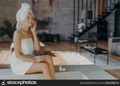Satisfied female model applies cosmetic product for healthy skin after showering, uses moisturizer and has daily routine, wrapped in bath towel, poses against home interior. Skin care procedure