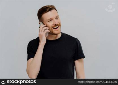 Satisfied ginger man talking on phone, receiving good news. Smiling, looking aside. Grey studio wall.