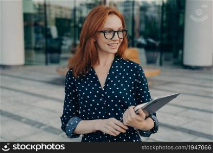 Satisfied ginger woman looks happily away wears spectacles polka dot dress carries laptop and organizer poses outside waits for someone near office building. People lifestyle technologies concept. Female model carries laptop and organizer poses outside waits for someone near office building