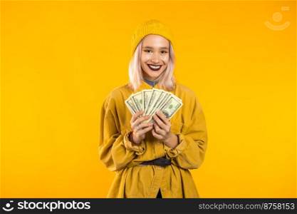 Satisfied happy excited unusual teen girl showing money - U.S. currency dollars banknotes on yellow wall. Symbol of success, gain, victory. Satisfied happy excited unusual teen girl showing money - U.S. currency dollars