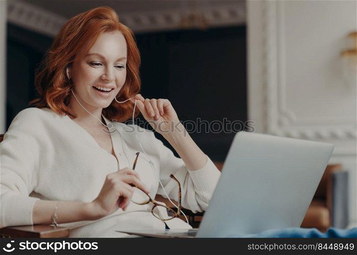 Satisfied happy foxy successful woman has video conference, uses modern laptop computer and earphones, browses data and works on remote project, poses in modern apartment, watches online webinar