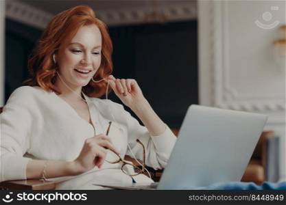Satisfied happy foxy successful woman has video conference, uses modern laptop computer and earphones, browses data and works on remote project, poses in modern apartment, watches online webinar