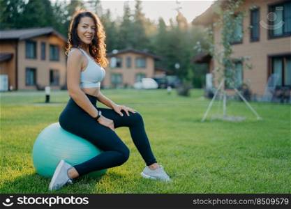 Satisfied healthy woman with curly dark hair sits on fitness ball, takes rest after yoga exercising, poses outdoor against private houses, enjoys spending free time on sport and favorite activity