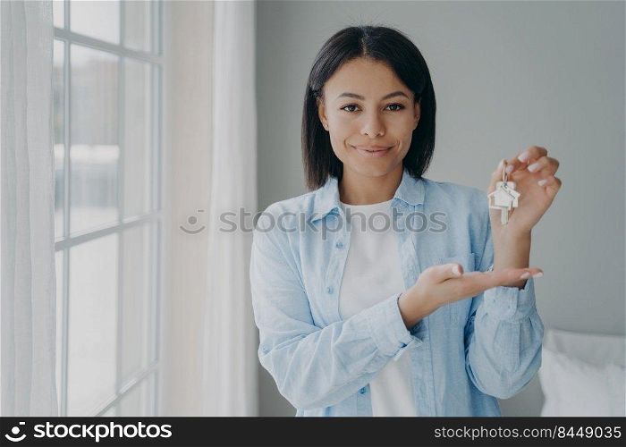 Satisfied hispanic girl is holding house keychain in her hand near window. Positive young woman is buying apartment and moving. Mortgage loan and investment concept. Modern scandinavian interior.. Satisfied hispanic girl holding house keychain in hand. Young woman is buying apartment and moving.