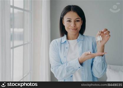 Satisfied hispanic girl is holding house keychain in her hand near window. Positive young woman is buying apartment and moving. Mortgage loan and investment concept. Modern scandinavian interior.. Satisfied hispanic girl holding house keychain in hand. Young woman is buying apartment and moving.
