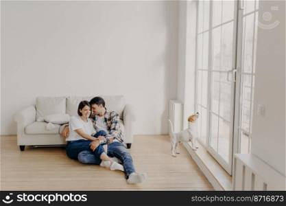 Satisfied husband and wife have romantic relationship, sit on floor near white couch in big room, wear jeans, tshirts and socks, spend leisure time in domestic atmosphere, dog poses near balcony
