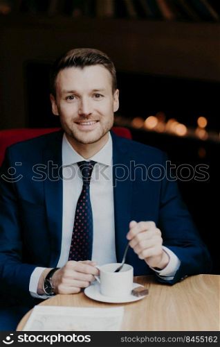 Satisfied male employee in luxury suit, drinks coffee, sits at wooden table in cafeteria, has pleasant smile and attractive look, waits for business partner or colleague going to discuss main problems