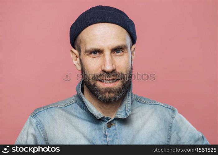 Satisfied male model with thick beard and mustache looks with confident expression into camera, wears fashionable hat and jacket, isolated over pink background. People, emotions, facial expressions