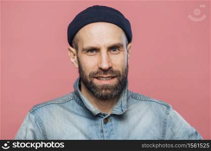 Satisfied male model with thick beard and mustache looks with confident expression into camera, wears fashionable hat and jacket, isolated over pink background. People, emotions, facial expressions