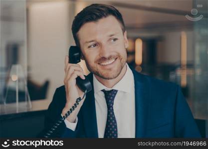 Satisfied male office worker calling his wife to notify her about promotion he just received, young handsome man with stubble and bright smile, office environment in blurred background. Satisfied male office worker calling his wife to notify her about promotion