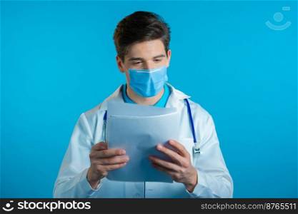 Satisfied man in professional medical coat and mask holding files papers isolated on blue background. He nods his head approvingly to diagnosis Doctor with stethoscope. High quality photo. Satisfied man in professional medical coat and mask holding files papers isolated on blue background. He nods his head approvingly to diagnosis Doctor with beard and stethoscope.