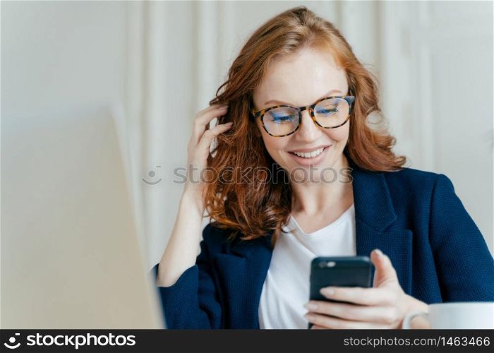Satisfied red haired female uses modern electronic device for checking newsfeed, online communication and searching information, sits in coworking space, uses high speed internet connection.
