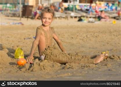 Satisfied six year old girl sitting on the beach and daubed themselves with wet sand