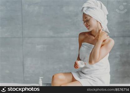 Satisfied spa woman sits in profile applies body cream for perfect skin, enjoys p&ering and beauty treatment poses wrapped in towel against grey wall in bathroom. Taking good care of own body
