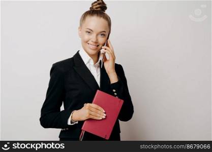 Satisfied woman in dark suit with notebook in hand talking on phone with partner about ideas or discussing working process, posing isolated on grey background. Business communication. Female business owner with phone and notebook