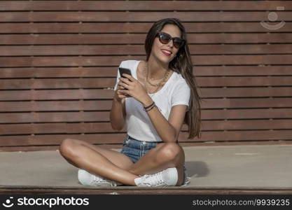 Satisfied woman in modern casual wear and sunglasses chilling on wooden bench with legs crossed and using smart phone in summertime. Smiling female browsing phone while relaxing in park