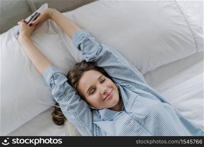 Satisfied woman stretches in bed, dressed in nightclothes, closes eyes with pleasure, holds cell phone, listens music in earphones, recreats in bedroom. People, good rest and modern technologies