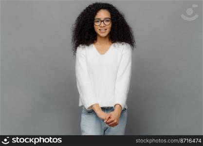 Satisfied young African American woman keeps hands together, smiles gently, dressed in casual white sweater and jeans, isolated over grey studio background. Pleasant emotions and feelings concept