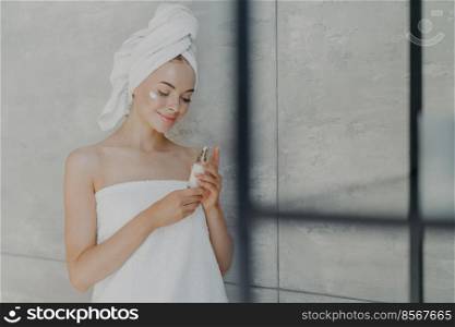 Satisfied young Caucasian woman cares of her beauty, applies lotion cream on face, stands wrapped in bath towel, poses in home bathroom against grey wall. Daily home care and day spa concept