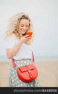 Satisfied young female with bushy curly hair holds smart phone, uses free internet connection for texting messages, stands against blurred seascape background. People and recreation concept.