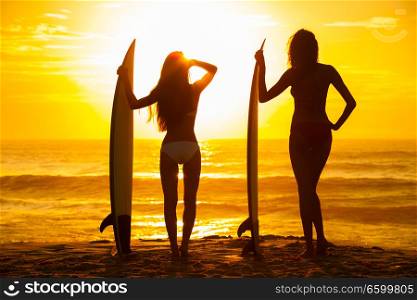 Saturated stylised rear view of two beautiful sexy young woman surfer girls in bikinis with white surfboards on a beach at sunset or sunrise
