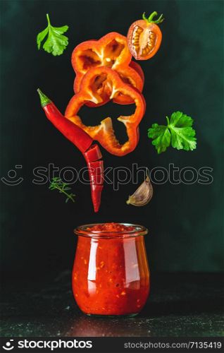Sauce components frozen in the air. Homemade DIY natural canned hot tomato sauce chutney with chilli or adjika in glass jar standing on wooden table with flying ingredients, selective focus