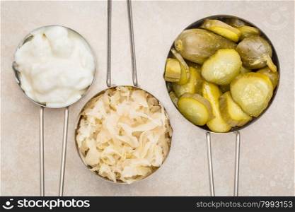 sauerkraut, cucumber pickles and yogurt - popular probiotic fermented food - three measuring cups against ceramic tile