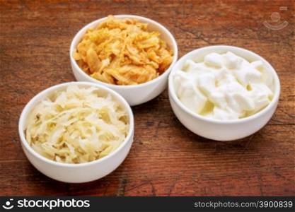 sauerkraut, kimchi and yogurt - popular probiotic fermented food - three ceramic bowl against rustic wood