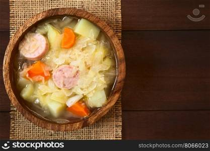 Sauerkraut soup or stew prepared with potato, carrot and bratwurst served in wooden bowl, photographed overhead on dark wood with natural light (Selective Focus, Focus on the top of the dish)