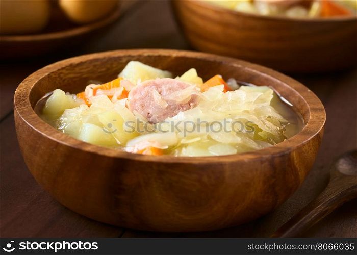 Sauerkraut soup or stew prepared with potato, carrot and bratwurst served in wooden bowl, photographed with natural light (Selective Focus, Focus in the middle of the dish)