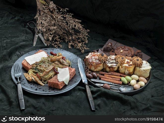 Sausage spaghetti with Cheese on bread in black ceramic plate served with Cinnamon Rolls, Dark chocolate bars, cookies and pieces in black ceramic plate on dark background. Space for text, Selective Focus.
