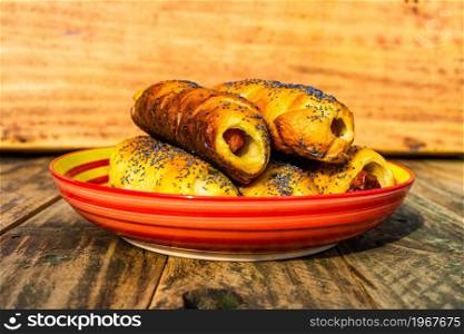 Sausages baked in dough sprinkled with salt and poppy seeds in a colorful plate. Sausages rolls, delicious homemade pastries in a rustic composition.. Sausages baked in dough sprinkled with salt and poppy seeds in a colorful plate. Sausages rolls, delicious homemade pastries in a rustic composition.