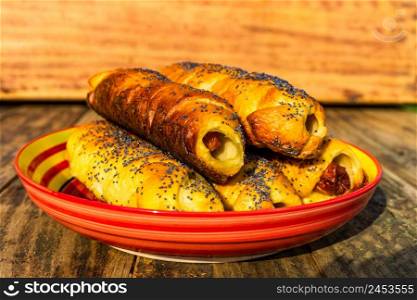 Sausages baked in dough sprinkled with salt and poppy seeds in a colorful plate. Sausages rolls, delicious homemade pastries in a rustic composition.. Sausages baked in dough sprinkled with salt and poppy seeds in a colorful plate. Sausages rolls, delicious homemade pastries in a rustic composition.