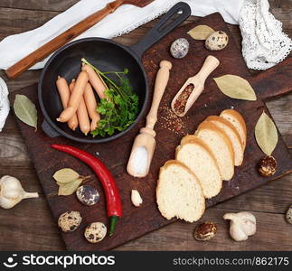 sausages in a black round cast-iron frying pan and raw quail eggs on a gray wooden table, top view