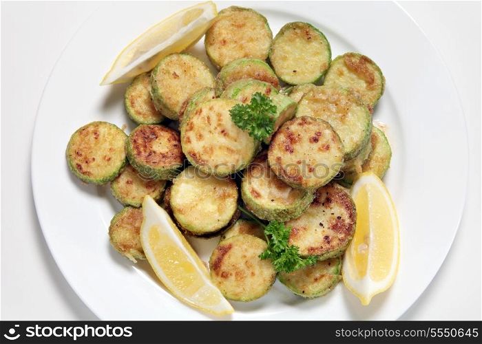 Sauteed courgette (zucchini) slices served with lemon wedges and garnished with parsley viewed from above. The courgettes are sliced, tossed in flour and fried in butter or olive oil and then seasoned.