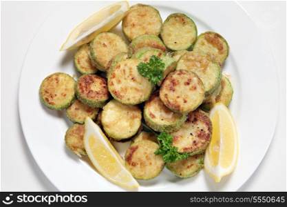 Sauteed courgette (zucchini) slices served with lemon wedges and garnished with parsley viewed from above. The courgettes are sliced, tossed in flour and fried in butter or olive oil and then seasoned.