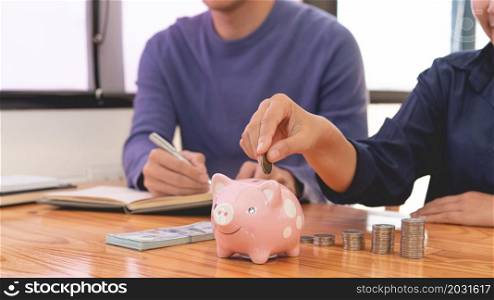 saving concept the man in blue sweater writing income account while the female in dark blue shirt drop a coin into a piggy bank.