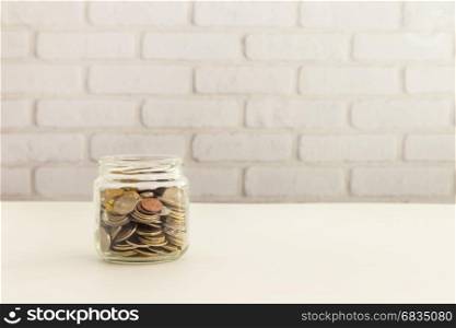 Saving money jar, world coins in glass container with white bricks wall background.