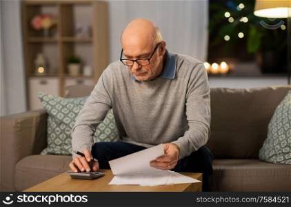 savings, annuity insurance and people concept - senior man with papers or bills counting on calculator at home in evening. old man with bills counting on calculator at home