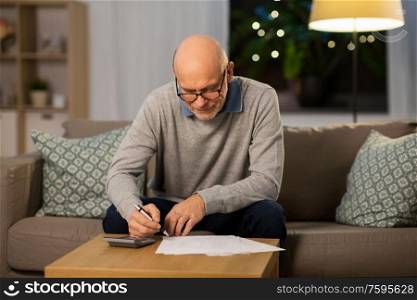 savings, annuity insurance and people concept - senior man with papers or bills and calculator writing at home in evening. senior man with bills and calculator at home