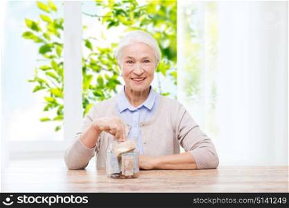 savings, annuity insurance and people concept - smiling senior woman putting money into glass jar at home over window and green natural background. senior woman putting money into glass jar at home